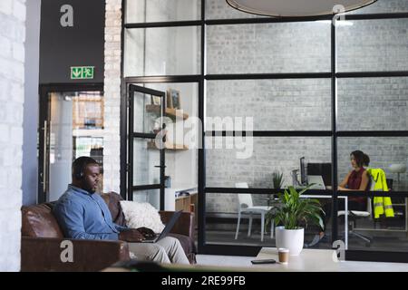Focused african american casual businessman sitting on couch and using laptop with headphones Stock Photo