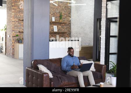 Focused african american casual businessman sitting on couch and using laptop with headphones Stock Photo