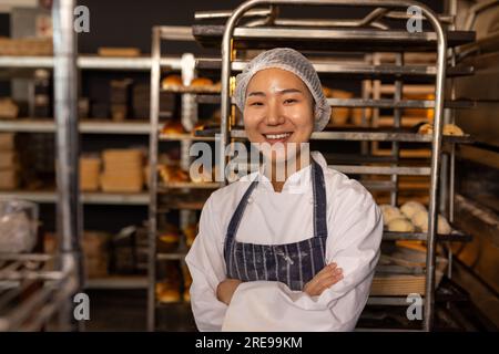 Portrait of happy asian female baker in bakery kitchen wearing apron with arms crossed Stock Photo