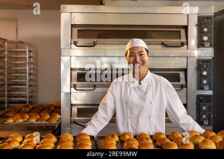 Portrait of happy asian female baker in bakery kitchen with fresh rolls Stock Photo