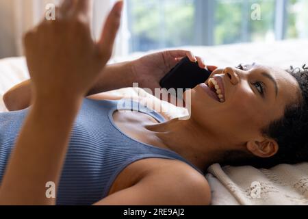 Happy biracial woman lying on bed and talking on smartphone in bedroom Stock Photo