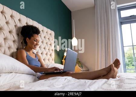 Happy biracial woman sitting on bed and using laptop in bedroom Stock Photo