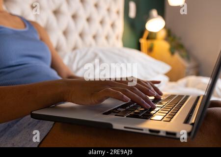 Midsection of biracial woman sitting on bed and using laptop in bedroom Stock Photo