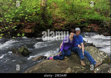 Young-at-heart couple rest on a boulder while hiking around Powhatan Lake near Asheville, North Carolina.  Both are wearing jeans, tennis shoes and sm Stock Photo