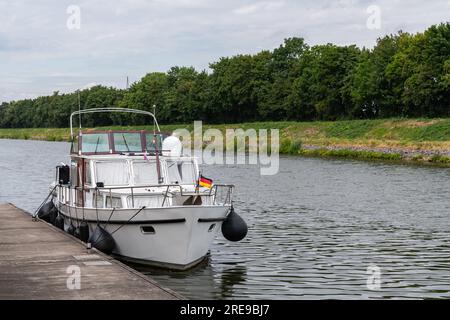 A small boat with a German flag on the bow is moored to a pier on the river. Stock Photo