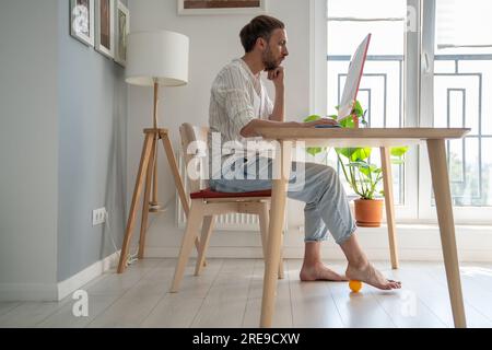 Man freelancer working on computer at home doing recovery foot exercise using massage silicone ball. Stock Photo