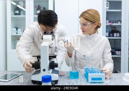 A team of scientists and chemists work in the laboratory on the study of substances, make analyses. An Asian man examines under a microscope, a woman fills flasks with blue liquid. Stock Photo