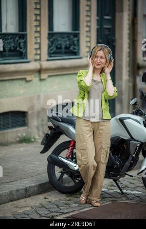 A woman listens to music with headphones while standing near a motorcycle on the street. Stock Photo