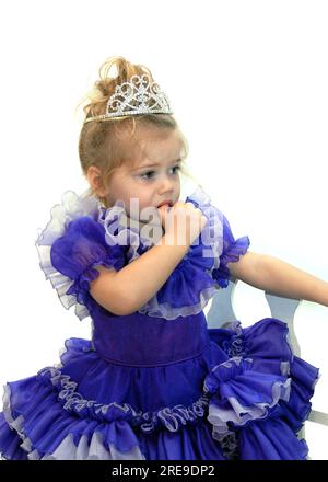 Little pageant girl fearfully sucks her thumb.  She is wearing a tiara and wearing a fancy purple pageant dress. Stock Photo