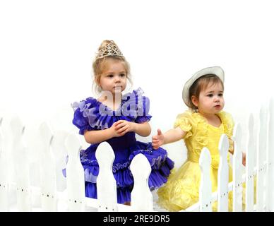 Two adorable little girls stand behind a white picket fence.  They are wearing pageant dresses in yellow and purple.  One has on a tiara the other a h Stock Photo