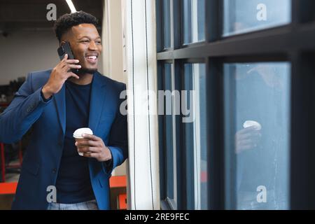 Happy african american man talking on smartphone while looking out of the window at office Stock Photo