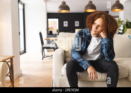 Thoughtful, sad biracial man sitting on couch in living room, looking away, copy space Stock Photo