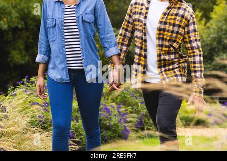 Midsection of diverse couple holding hands, walking in garden Stock Photo