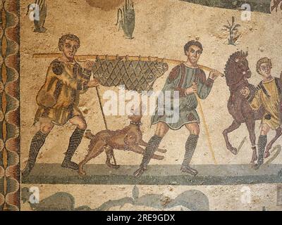 Mosaic in the Room of the Small Hunt, Villa Romana del Casale, Sicily, depicts two men with a hunting dog carrying a trussed boar on a pole. Stock Photo