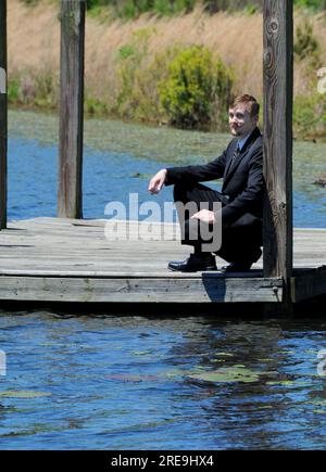 Handsome young man relaxes on a wooden dock.  Lake water is blue and dock is rustic.  He is wearing a black business suit. Stock Photo