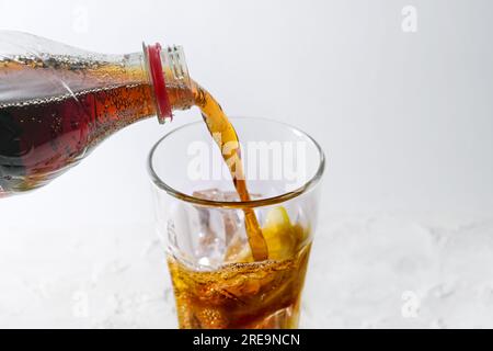 Pouring fresh cola from a bottle into a drinking glass with ice cubes and lemon slice, refreshing drink against a light gray background, copy space, s Stock Photo