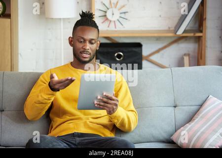 Young african man working remotely, having line video call with team, sitting on sofa at home. Online meeting Stock Photo