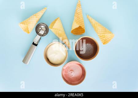 Ice cream in a box with different flavors stands on a blue background along with a metal ice cream scoop and a waffle cone. Selective focus, noise Stock Photo