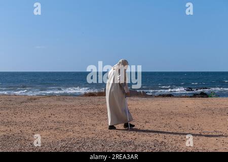 A picture from behind of an old woman with crutch walking on the seashore, the blue sea is in a pleasant atmosphere, Old woman in robes on the beach Stock Photo