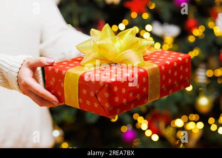 Womans hands in cozy sweater holding present with golden ribbon in festive room Stock Photo