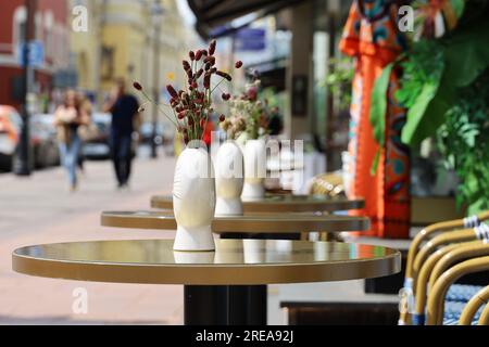 Street cafe in city with empty tables outdoor on walking people background. Vases of flowers on round tables and cozy chairs Stock Photo