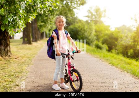 Cheerful girl arriving at school on her bike, wearing backpack for the start of school, standing near bicycle in park Stock Photo