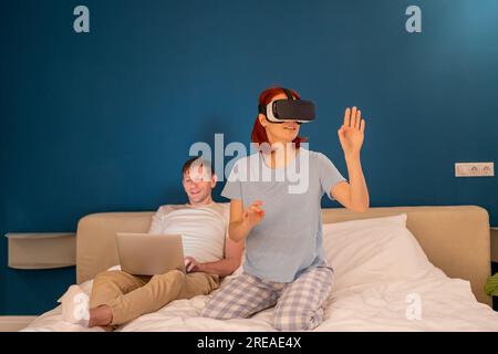 Interested woman in glasses virtual reality sits on bed with husband manages vr goggles from laptop Stock Photo