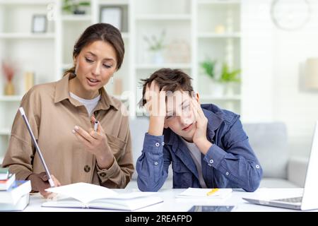 Cute and boring study at home, mom explains homework to son, family study at home sitting in living room, son doesn't understand homework upset and bored. Stock Photo