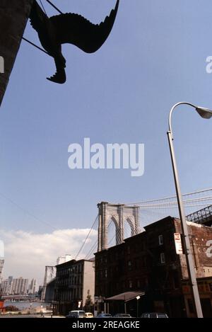 AJAXNETPHOTO. JULY, 1975. BROOKLYN, NEW YORK, USA. - EAGLE SCULPTURE - ICONIC BROOKLYN EAGLE LANDMARK ON OLD WHAREHOUSE BUILDING OVERLOOKS BROOKLYN BRIDGE TOWER EMERGING BEHIND NINETEENTH CENTURY COMMERCIAL BUILDINGS ON CADMAN PLAZA WEST BELOW THE DECK OF THE BROOKLYN BRIDGE SPANNING EAST RIVER BETWEEN PARK ROW MANHATTAN AND SANDS STREET, BROOKLYN, NEW YORK CiTY.   PHOTO:JONATHAN EASTLAND/AJAXREF:601874 140 2 Stock Photo