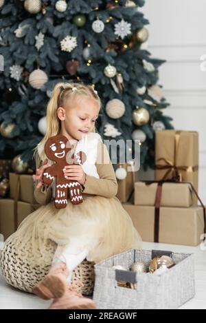 Merry Christmas and Happy Holidays. Child girl eating big iced ingerBread man cookies under Christmas tree Waiting for Christmas. Celebration New Year Stock Photo