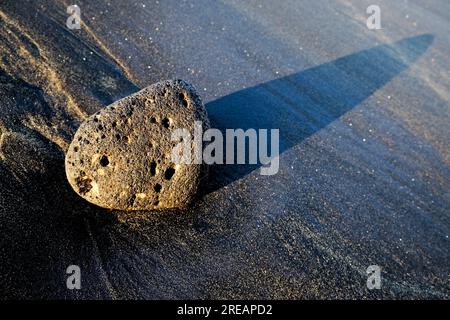 Large black basalt lava boulder at sunset on a black volcanic sand beach. Stock Photo