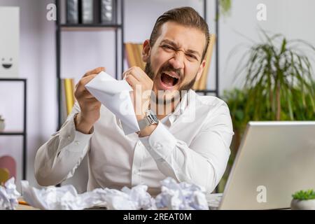 Angry furious businessman working at office throwing crumpled paper, having nervous breakdown at work, migaine, headache, stress management, mental distress problems, losing temper reaction on failure Stock Photo