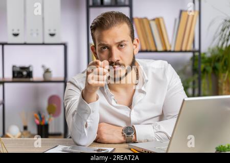 I choose you. Middle eastern businessman programmer software developer working on office laptop pointing to camera, looking with happy expression, making choice, showing direction. Arab freelancer man Stock Photo