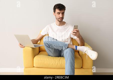 Young man with laptop using mobile phone in armchair near light wall Stock Photo