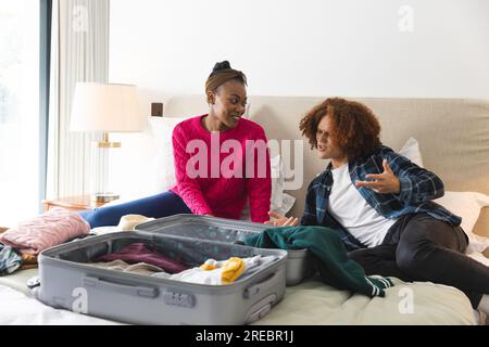 Happy diverse couple lying on bed, talking and packing suitcase in sunny bedroom, at home Stock Photo