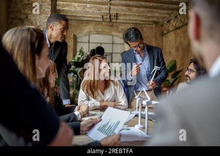 A young Indian engineer with glasses presents a wind turbine model in a multicultural office. Tablets, charts, and more models are on the shared desk. Stock Photo