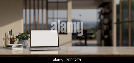 Close-up workspace with a laptop white screen mockup, books, painting tools, and a potted plant on a tabletop with a blurred modern office as a backgr Stock Photo