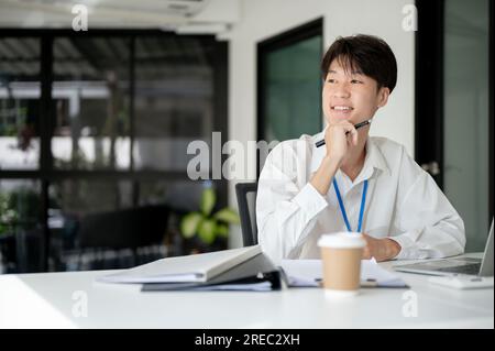 A smart and thoughtful young Asian male office worker is looking out the window and daydreaming about his career success while sitting at his desk in Stock Photo