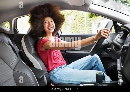 Side view of young African American female in casual clothes with Afro hairstyle sitting and laughing in car looking at camera Stock Photo