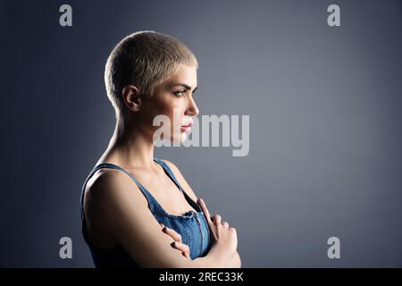 Side view of serious young female model with short blond hair standing arms crossed and looking away against gray background Stock Photo