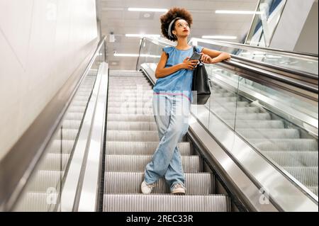 Low angle full length of pensive black woman looking up while standing on escalator in light subway Stock Photo