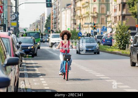 Happy African American female riding bicycle on asphalt cycle path near road with cars and looking at camera Stock Photo