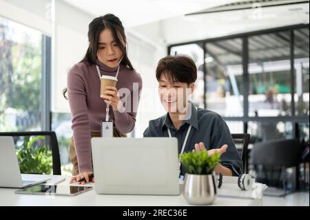 A professional and focused Asian female marketing manager is reading details on a laptop and helping a young male employee with a marketing campaign, Stock Photo