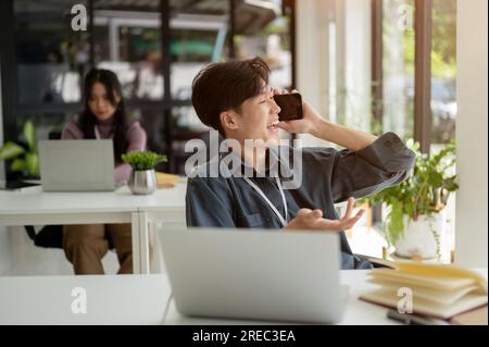 A cheerful young Asian businessman is having an informal phone call with his friend while sitting in his office during the coffee break. Stock Photo