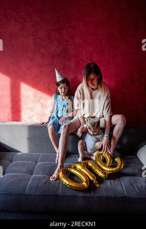 Tired young mother sits on sofa with small children. Red wall. Single parent family celebrating babys first year. Foil balloons ONE on gray couch. Lif Stock Photo