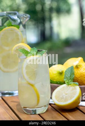 Lemonade in glass and pitcher with mint and sliced lemons outdoors on wooden patio table and nature background Stock Photo