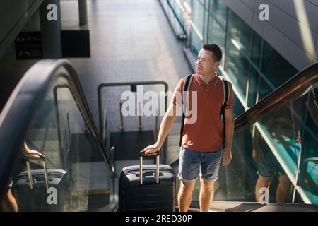 Traveler walking through airport terminal. Man with suitcase standing on ecalator. Passenger is ready for travel by airplane. Stock Photo