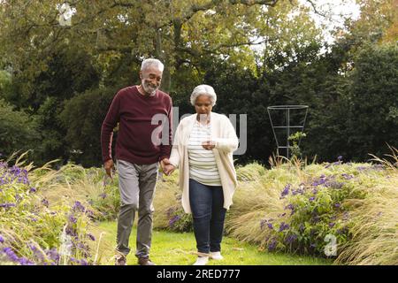 Happy senior biracial couple holding hands and walking in garden at home Stock Photo