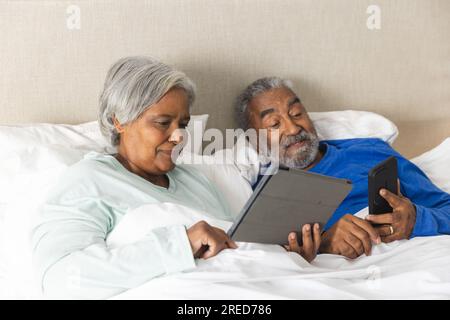 Happy senior biracial couple lying in bed and using tablet and smartphone at home Stock Photo