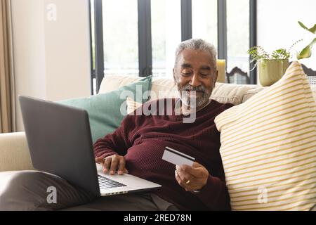 Happy senior biracial man sitting on couch using laptop and credit card at home Stock Photo